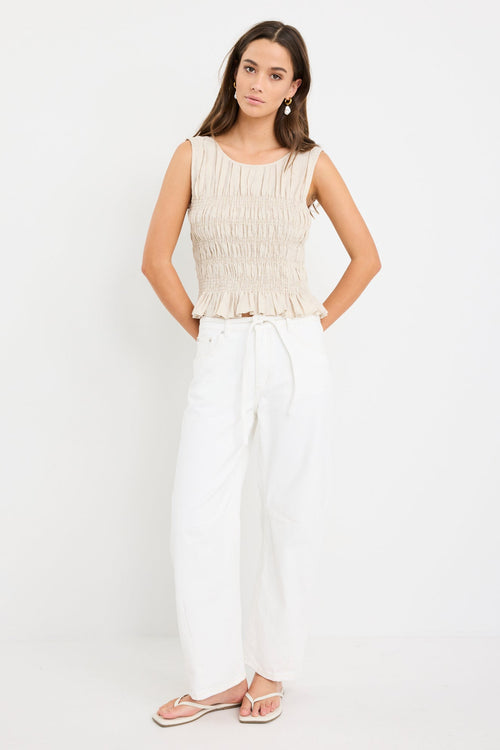 Woman wearing a natural linen sleeveless top with shirred bodice and ruffle hem, styled with ecru jeans.