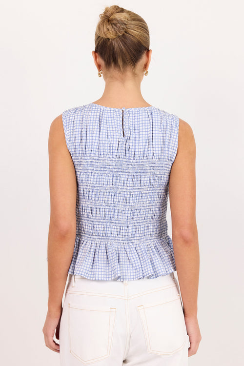 A woman viewed from the back wearing a sleeveless smocked top with a light blue and white gingham pattern and white pants, set against a plain white background.