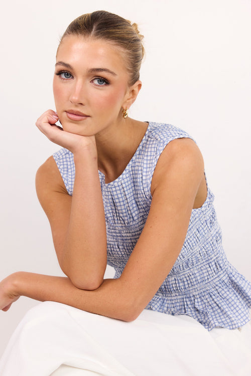 A woman wearing a blue and white gingham sleeveless top and white pants, posing with her hand on her chin against a plain white background.