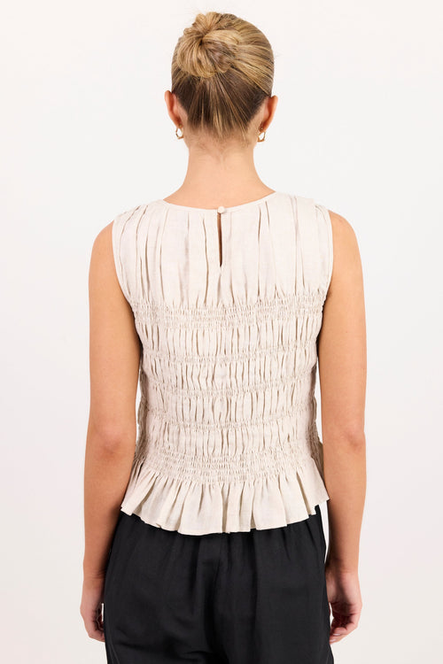 A woman viewed from the back wearing a sleeveless, off-white smocked peplum top and black pants, set against a plain white studio background.