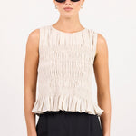 A model stands against a plain light gray studio background wearing a sleeveless cream pleated top with a ruffled hem and black wide-leg trousers.