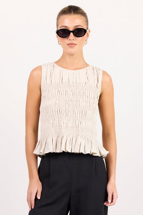 A model stands against a plain light gray studio background wearing a sleeveless cream pleated top with a ruffled hem and black wide-leg trousers.