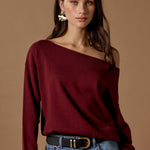 Woman wearing a burgundy off-shoulder top and blue jeans against a brown background