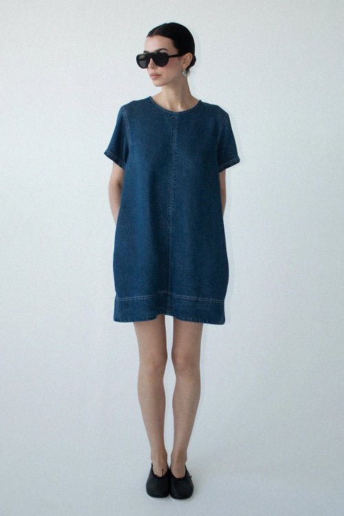 A woman modeling a dark indigo denim short-sleeve tunic dress with a raw hem, styled with black sunglasses and black slip-on shoes against a plain light gray background.