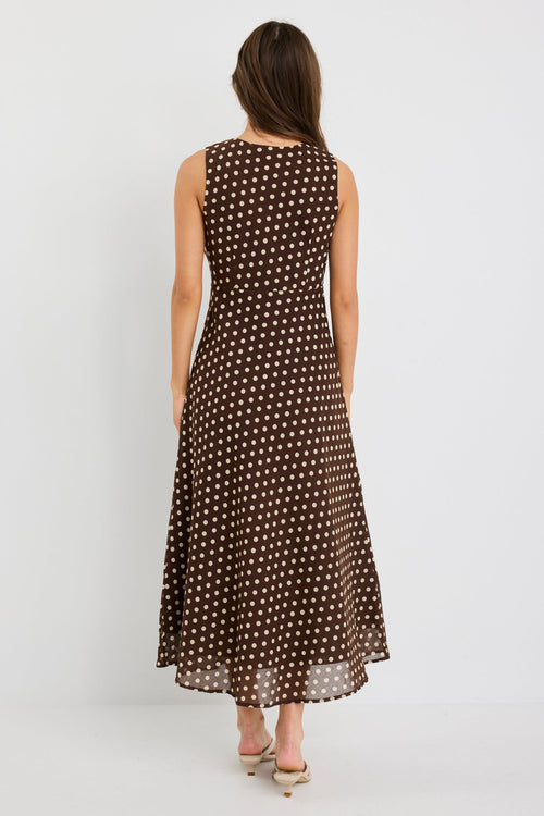 Woman wearing a sleeveless chocolate brown bias-cut midi dress with ivory polka dots, V-neckline and tie detail, styled with neutral sandals.
