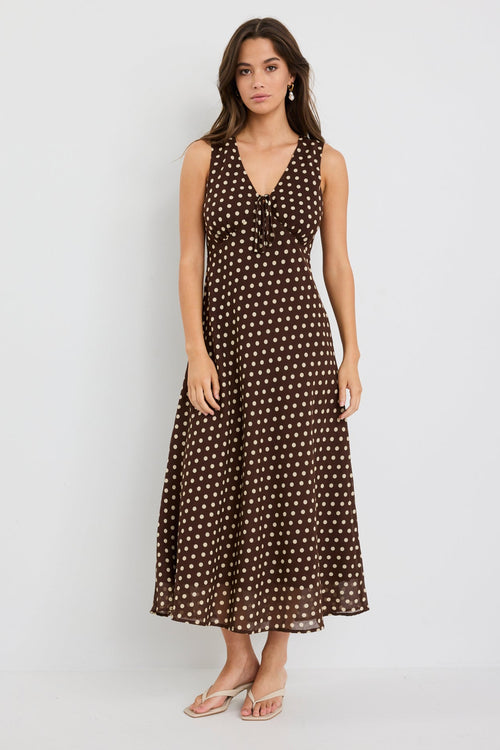 Woman wearing a sleeveless chocolate brown bias-cut midi dress with ivory polka dots, V-neckline and tie detail, styled with neutral sandals.