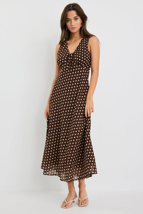 Woman wearing a sleeveless chocolate brown bias-cut midi dress with ivory polka dots, V-neckline and tie detail, styled with neutral sandals.