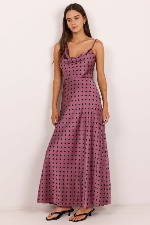 A woman modeling a sleeveless, floor-length cowl-neck dress with a dusty rose base and black polka dots, paired with black flat sandals, set against a plain off-white studio background.