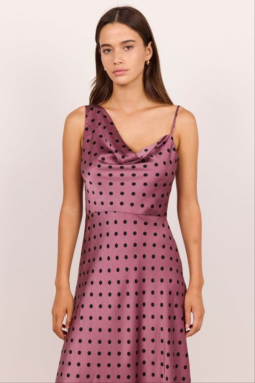A woman modeling a sleeveless cowl-neck midi dress with thin spaghetti straps and a black polka dot pattern on a dusty rose pink background, set against a plain light beige studio backdrop.