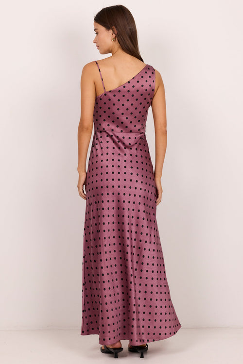 A woman modeling a one-shoulder maxi dress with a black polka dot pattern on a dusty purple background, shown from the back against a plain white studio backdrop.