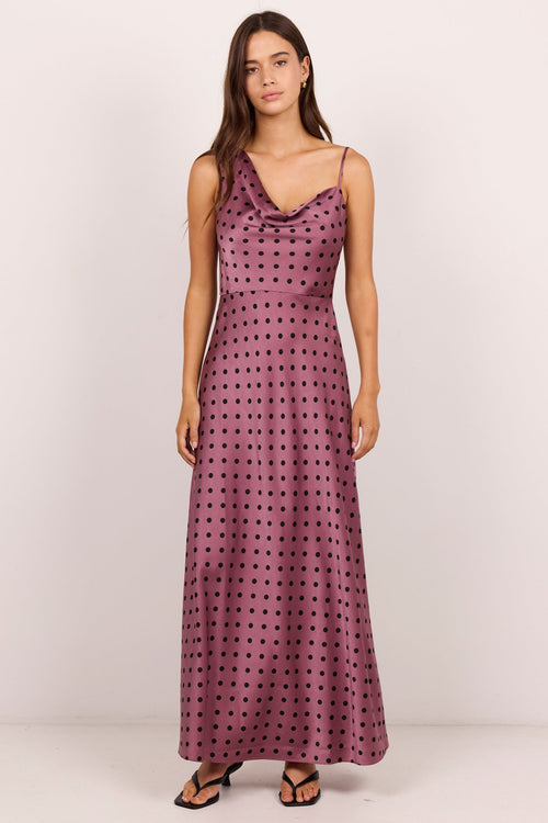 A woman modeling a sleeveless, floor-length mauve maxi dress with a black polka dot pattern, standing against a plain off-white studio background.