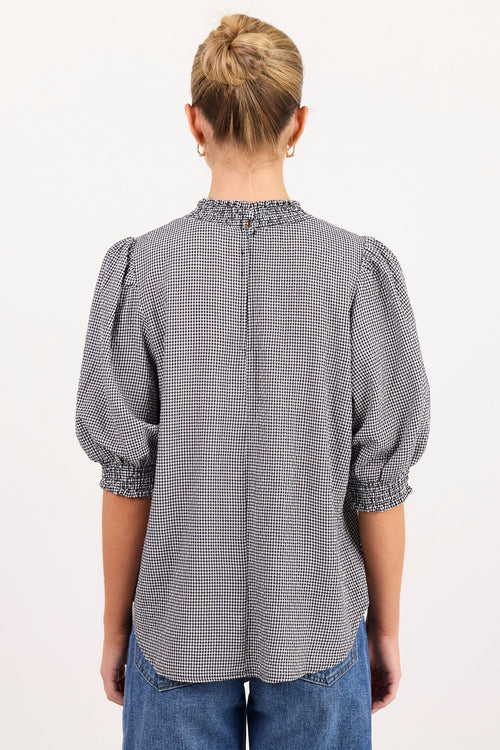 A woman viewed from behind wearing a gray and white micro-check patterned blouse with puffed elbow-length sleeves and a smocked neckline, paired with blue jeans against a plain white background.