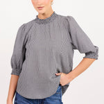 A woman modeling a gray and white micro-checkered blouse with a textured embroidered yoke and rolled three-quarter sleeves, paired with blue jeans against a plain white studio background.