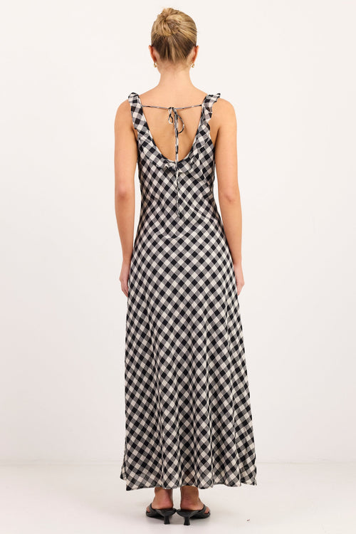 A woman viewed from behind wearing a sleeveless black and white gingham maxi dress with tie straps at the back, standing against a plain white studio background.