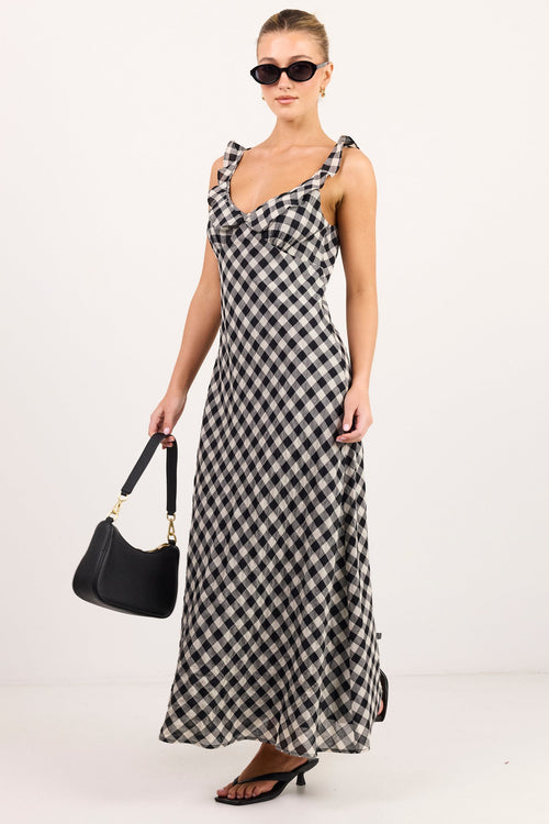 A woman modeling a sleeveless black and white gingham maxi dress with a V-neckline, holding a black handbag, set against a plain white studio background.