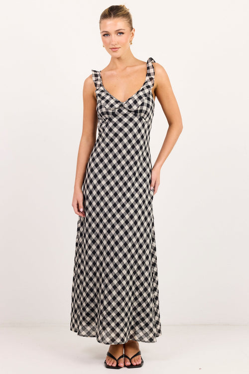 A woman modeling a sleeveless black and white gingham maxi dress with shoulder tie details, standing against a plain white studio background.