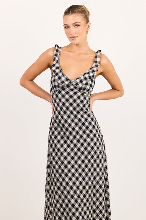 A woman modeling a sleeveless black and white gingham maxi dress with a wrap-style V-neckline, posed against a plain white studio background.