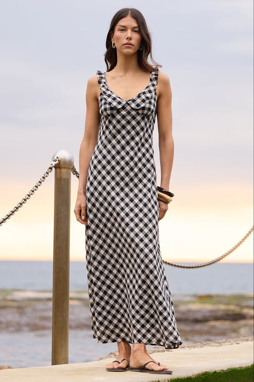 A woman stands on a concrete pier by the ocean wearing a black and white checkered sleeveless maxi dress. The background features a calm sea, rocky shoreline, and a cloudy sky with a soft sunset glow.
