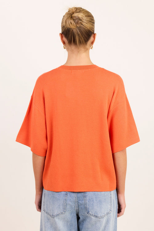 A person viewed from the back wearing a loose orange short-sleeve T-shirt and light blue jeans against a plain light background.