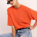 A woman wearing an oversized orange short-sleeve crewneck t-shirt and light-wash blue jeans, sitting against a plain off-white background.