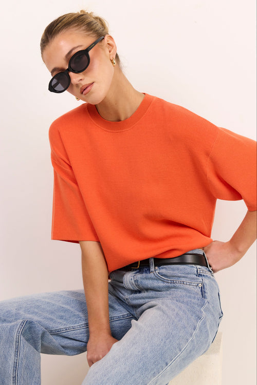 A woman wearing an oversized orange short-sleeve crewneck t-shirt and light-wash blue jeans, sitting against a plain off-white background.