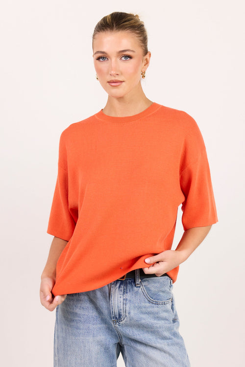 A woman modeling an oversized short-sleeve crewneck t-shirt in a vibrant orange color, paired with light-wash blue jeans, against a plain light beige studio background.