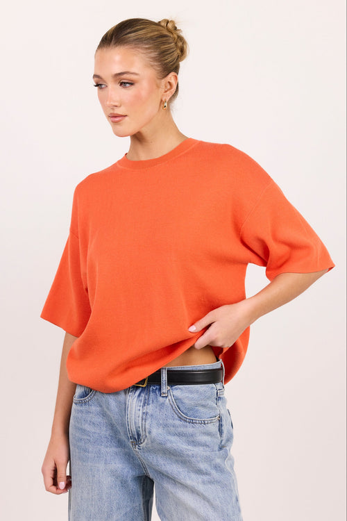 A woman wearing a loose, short-sleeve crewneck sweater in a vibrant orange, paired with light-wash blue jeans and a black belt, photographed against a solid off-white studio background.
