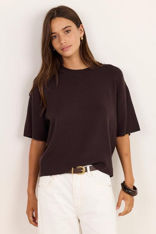 A woman modeling a dark brown short-sleeve crewneck t-shirt paired with white pants and a gold belt, standing against a plain off-white wall.