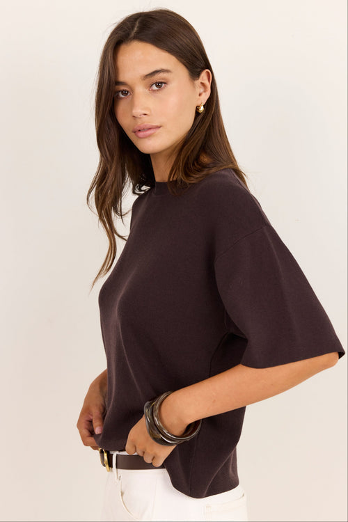 A woman modeling a dark brown short-sleeve knit top with a crew neck, paired with white pants, against a plain off-white background.