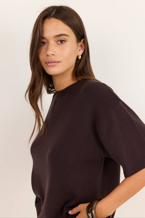 A woman wearing a dark brown short-sleeve crewneck knit top, photographed against a clean off-white studio background.