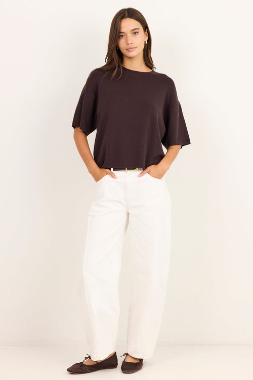 A woman modeling a dark brown short-sleeve top and white wide-leg pants against a plain white studio background.