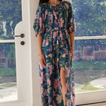 A woman modeling a dark teal maxi dress with a pink and green tropical floral print, standing in a bright doorway with a garden view.