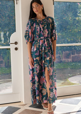 A woman models a floral midi dress with a high slit, standing in a doorway with a garden view in the background. The dress features a dark teal base with pink, purple, and green tropical floral patterns.