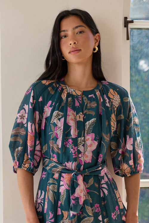 A woman wearing a dark teal floral midi dress with puff sleeves and a self-tie waist, standing in front of a white arched doorway with greenery visible outside.