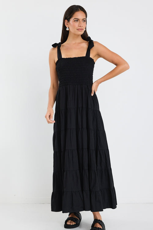 Woman wearing a black tiered maxi dress with shirred bodice and bow-tie straps, styled with statement earrings and woven black slides.