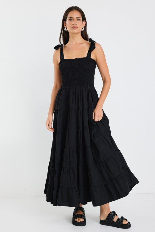 Woman wearing a black tiered maxi dress with shirred bodice and bow-tie straps, styled with statement earrings and woven black slides.