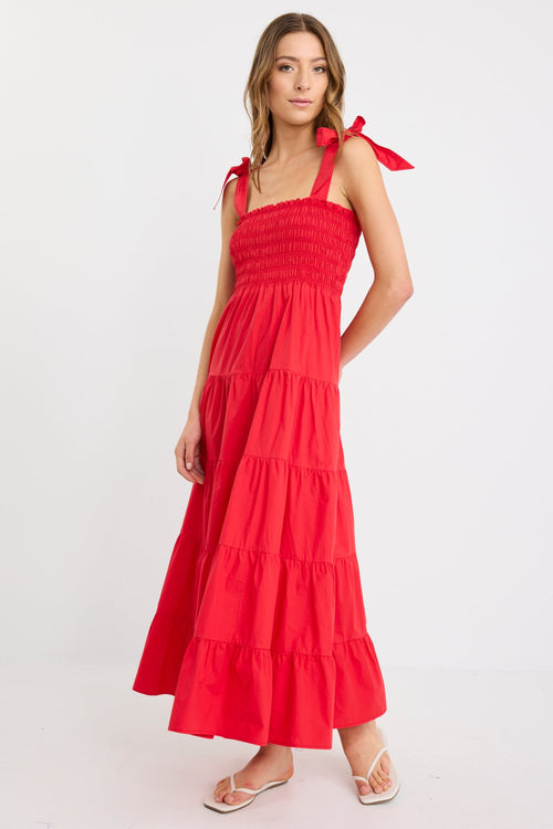 Woman wearing a cherry red tiered maxi dress with shirred bodice and strappy tie shoulders, styled with flat sandals.