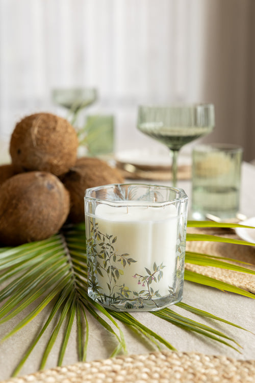 A white candle in a clear glass holder with a delicate green botanical leaf pattern, styled on a table with palm leaves, coconuts, and green glassware in the background.