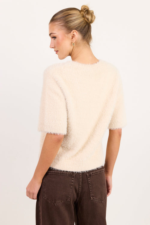 A woman shown from the back wearing a cream-colored, short-sleeved fuzzy knit sweater and dark brown pants against a plain white studio background.