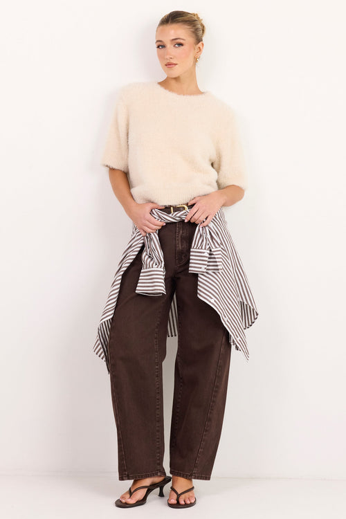 A woman stands against a plain white studio wall wearing a cream short-sleeve knit top, dark brown wide-leg pants, and a black-and-white striped shirt tied around her waist.