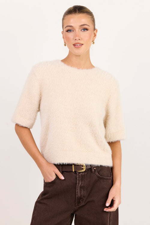 A woman modeling a short-sleeved, fuzzy knit sweater in cream, paired with dark brown wide-leg jeans and a gold-buckled belt, against a plain white studio background.