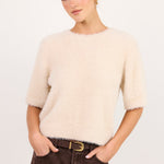 A woman modeling a short-sleeved, fuzzy cream-colored knit sweater paired with dark brown wide-leg jeans and a gold-buckled belt, set against a plain light gray studio background.