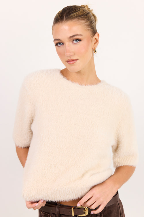 A woman wearing a fuzzy short-sleeve knit sweater in cream, paired with dark brown trousers and a gold-buckled belt, against a plain white studio background.