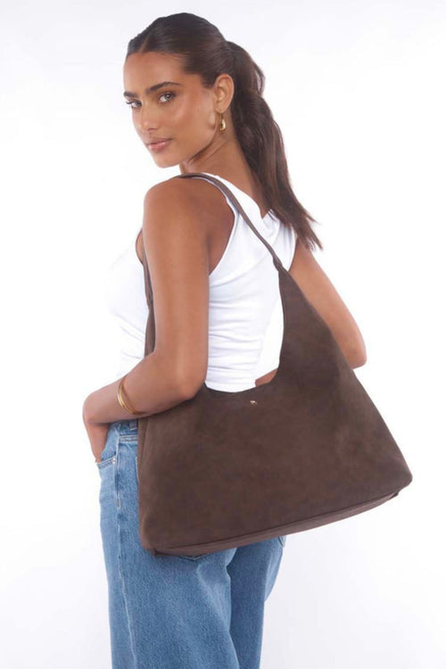 Woman wearing a white tank top and jeans, carrying a chocolate brown suede shoulder bag with a slouchy silhouette.