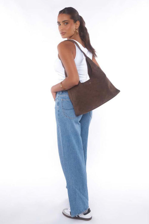 Woman wearing a white tank top and jeans, carrying a chocolate brown suede shoulder bag with a slouchy silhouette.