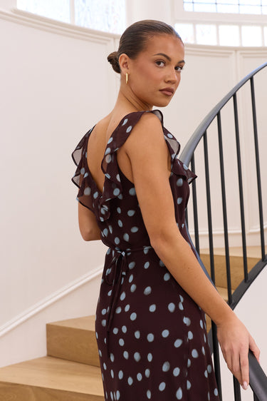 A woman wearing a sleeveless dark plum dress with light blue polka dots and ruffled shoulder details, posing on a wooden staircase next to a black metal spiral railing in a bright, modern interior.