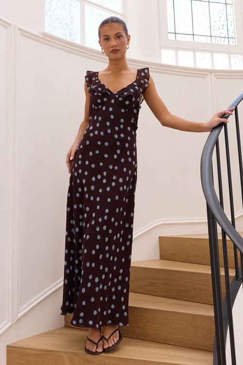 A woman wearing a sleeveless dark brown maxi dress with a light blue polka dot pattern stands on wooden stairs inside a bright white building, holding onto a black metal railing.