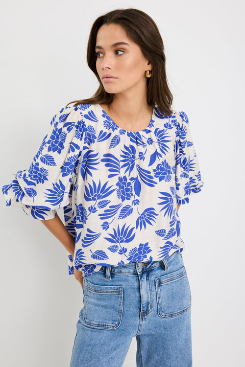 Woman wearing a cream blouse with bold blue floral print and blouson short sleeves, styled with light blue denim jeans.