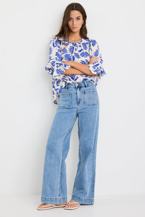 Woman wearing a cream blouse with bold blue floral print and blouson short sleeves, styled with light blue denim jeans.