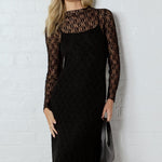Woman wearing a black dress with sheer sleeves against a white brick wall.
